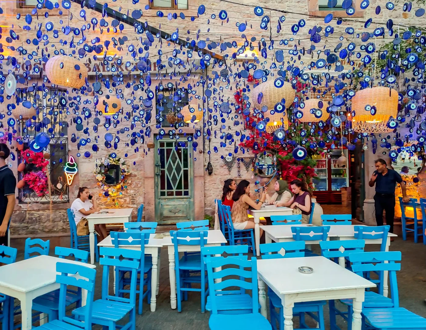 Cunda Adası'nın taş sokaklarından birinde yer alan, tavandan sarkan mavi nazar boncukları ve hasır lambalarla hareketlenmiş bu açık hava Ege kafesinde, mavi ahşap sandalyelerin ve beyaz masaların etrafında oturan insanlar keyifli bir road trip molası veriyor.