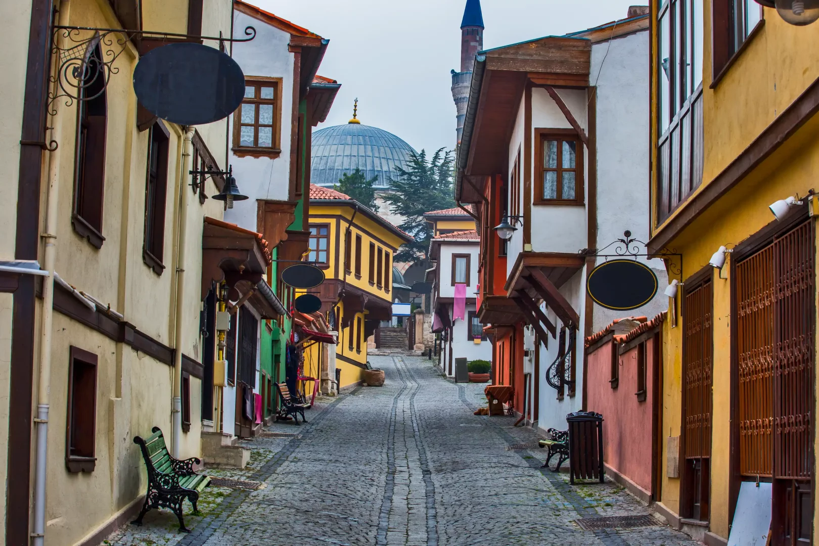 Eskişehir Odunpazarı'nın Arnavut kaldırımlı dar sokağında karşılıklı sıralanmış, renkli ahşap cumbalı tarihi Osmanlı evleri ve gerideki cami kubbesi, Türkiye road trip rotalarının kültürel zenginliğini vurguluyor.