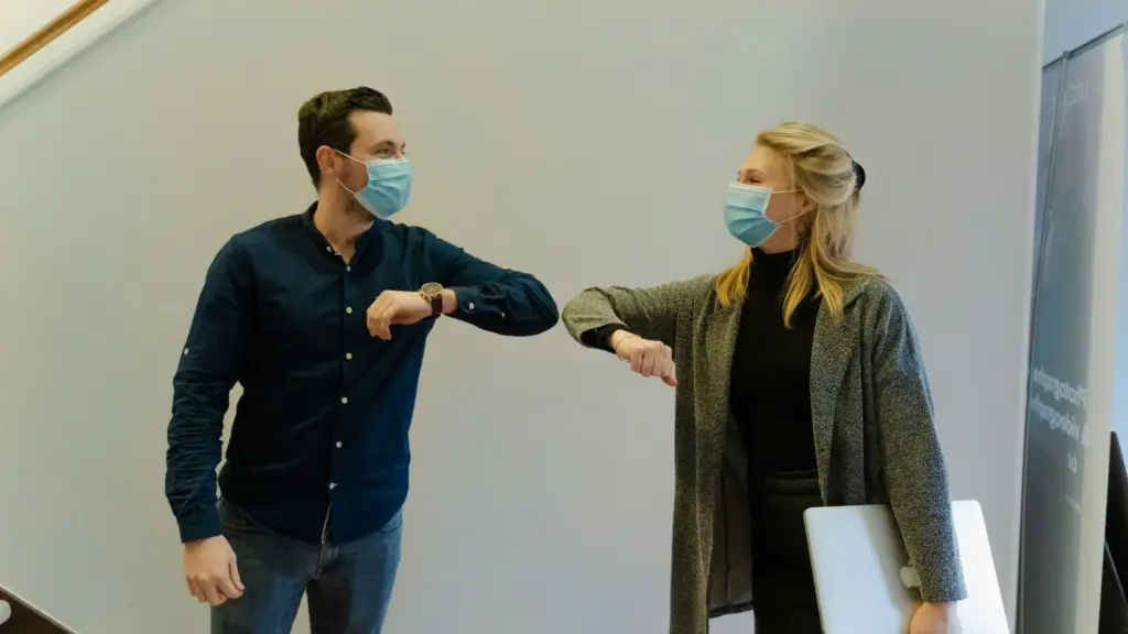 Maske takan iki kişi dirsek temasıyla selamlaşırken, COVID-19 döneminde sosyal mesafe ve hijyen kurallarına uygun karşılaşma.