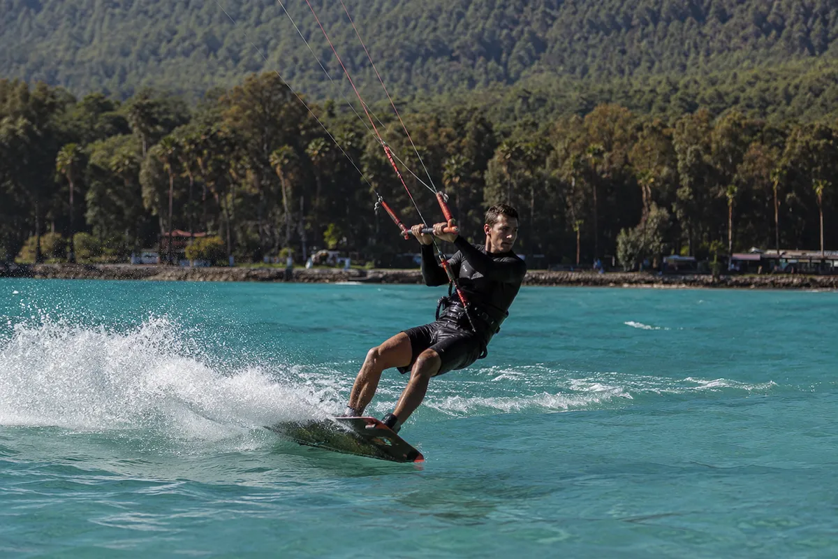 Akyaka'nın berrak turkuaz sularında, arkasında bolca su sıçratarak sörf tahtası üzerinde dengede duran ve kontrol barını ustaca tutan dalış kıyafeti giymiş genç bir adam kitesurf yaparken, arka planda yemyeşil ağaçlarla kaplı dağlar ve kıyı şeridi görülüyor.