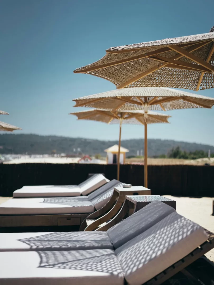 Çeşme'deki bir beach club'da, açık mavi gökyüzünün altında, hasır şemsiyelerin oluşturduğu dantel gibi gölgelerin düştüğü konforlu gri şezlonglar kumların üzerinde sıralanmış, dinlenmek ve güneşlenmek için ideal bir ortam sunuyor.