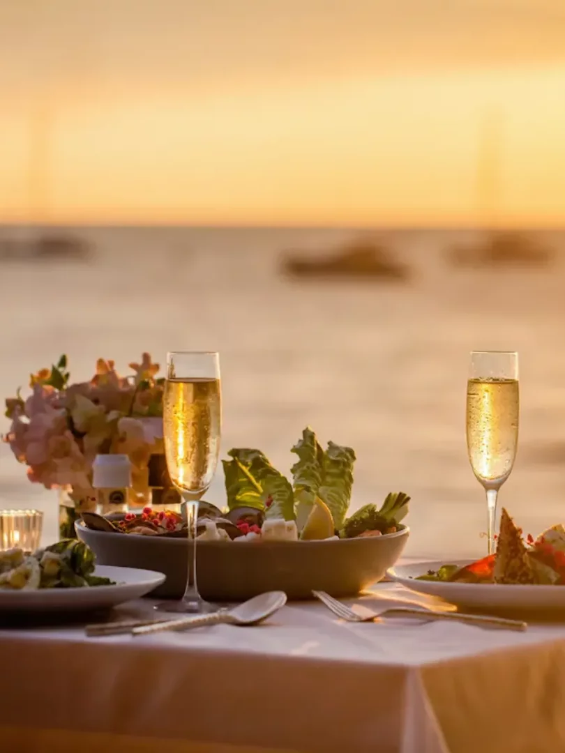 Çeşme'deki en iyi restoranlardan birinde denize nazır, gün batımının romantik atmosferinde, iki kadeh köpüklü şarap, zengin salata ve özenle hazırlanmış ana yemeklerle donatılmış beyaz örtülü şık bir masanın görünümü.