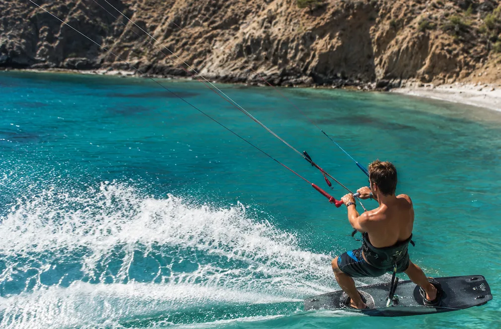 Masmavi berrak sularda, rüzgarın gücüyle bir sörf tahtası üzerinde ilerleyen ve geride köpüklü bir iz bırakan bir adamın Kite Surf yaptığı an; uzak arka planda ise sakin bir koyu çevreleyen görkemli kayalıklar ve kumsal yer alıyor.