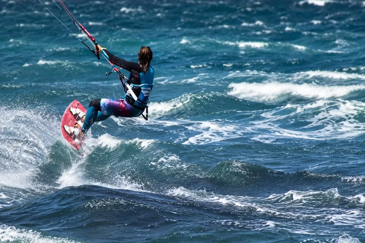 Gökçeada ve Çanakkale'de kitesurf yapılabilecek dalgalı deniz koşullarını gözler önüne seren bu dinamik fotoğrafta, rengarenk wetsuit giymiş bir kitesurfçü, kırmızı kitesurf tahtasıyla mavinin farklı tonlarındaki dalgaların üzerinde zıplayarak havada akrobatik bir hareket gerçekleştiriyor.