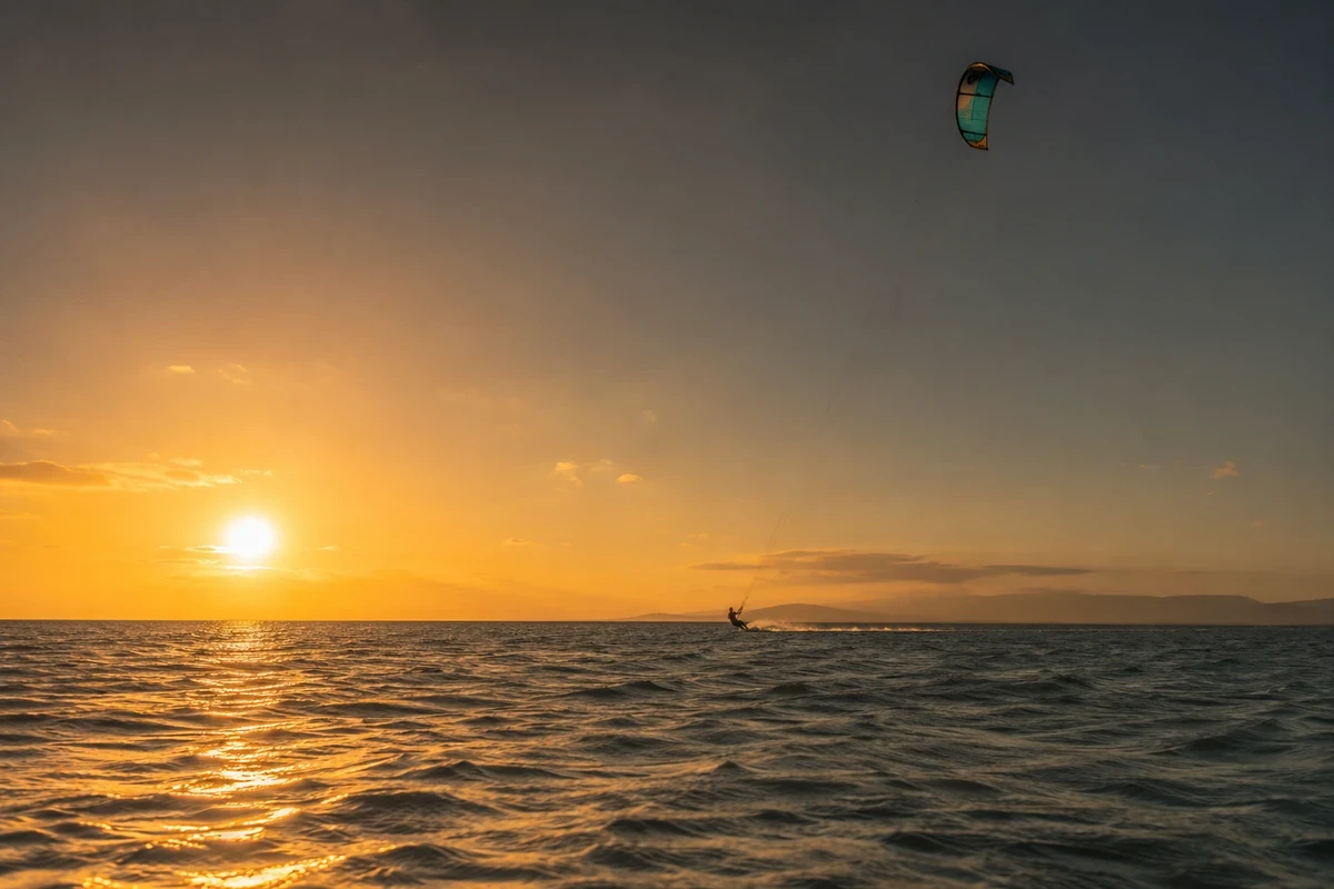 Geniş açıyla çekilmiş bu etkileyici gün batımı manzarasında, ufukta batmakta olan güneşin turuncu ve altın ışıkları denize yansırken, renkli kitesurf uçurtmasıyla bir sporcu dalgalar üzerinde hızla süzülerek arkasında beyaz bir köpük izi bırakıyor.