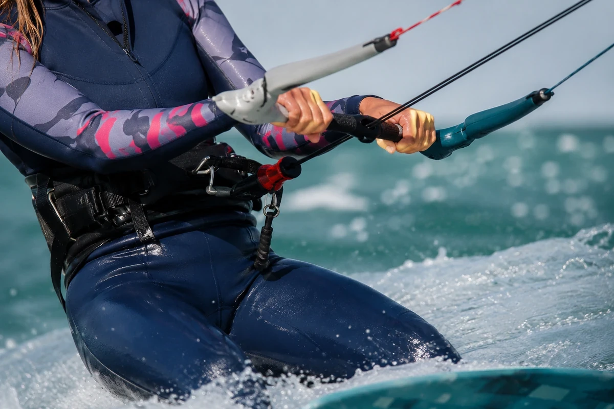 Koyusu gri-pembe desenli neopren elbise giymiş, bel kuşağı takan bir kitesörfçünün, köpüklü su üzerinde kitesurf kontrol barını sıkıca kavrayan elleri ve ıslak kolları detaylıca gösteriliyor.