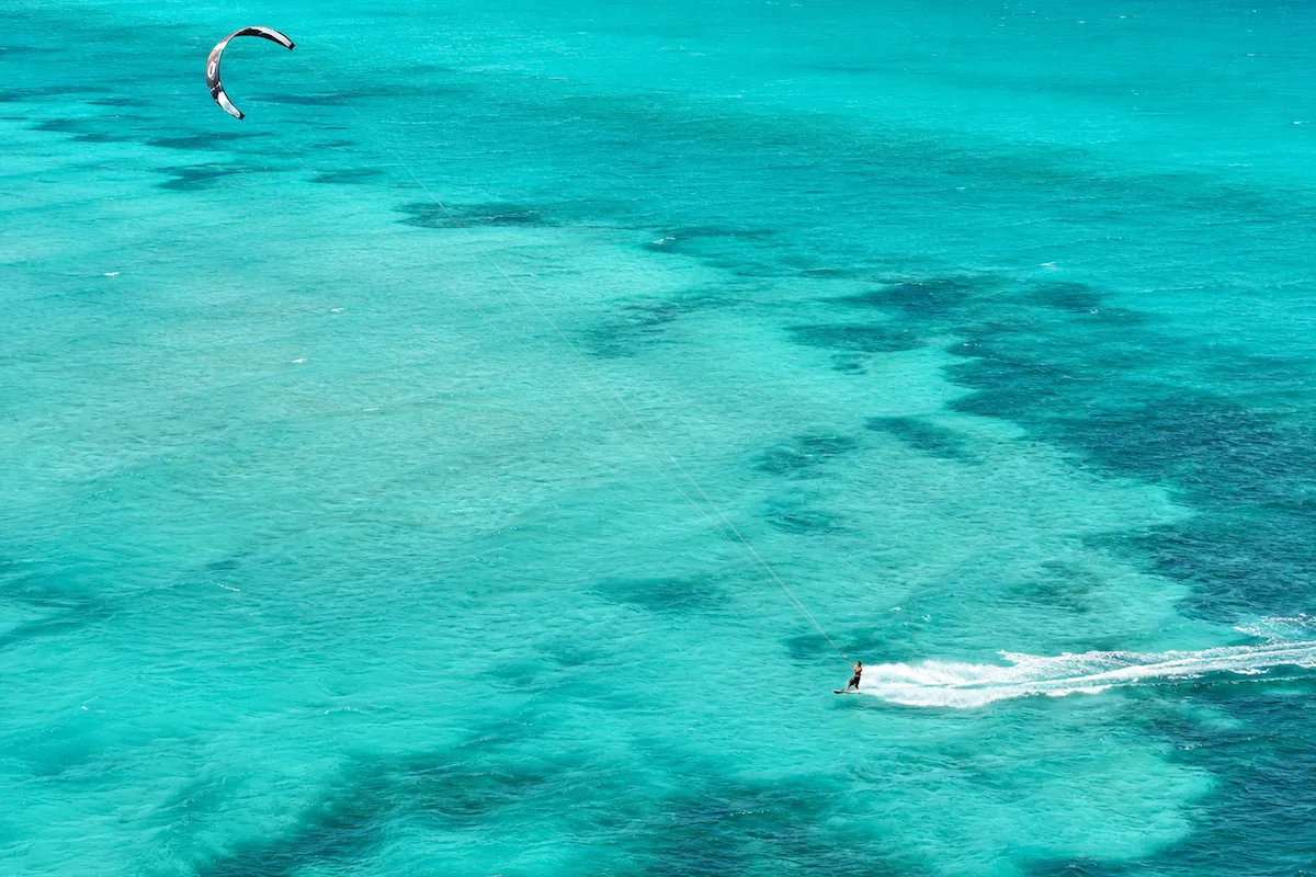 Urla'nın kristal berraklığındaki turkuaz sularında, rüzgarın gücüyle hızla ilerleyen Kitesurf yapan insan, su üzerinde büyüleyici beyaz bir köpük izi bırakarak kayıyor ve üstünde siyah, beyaz ve kırmızı detaylara sahip uçurtması gökyüzünde süzülüyor.