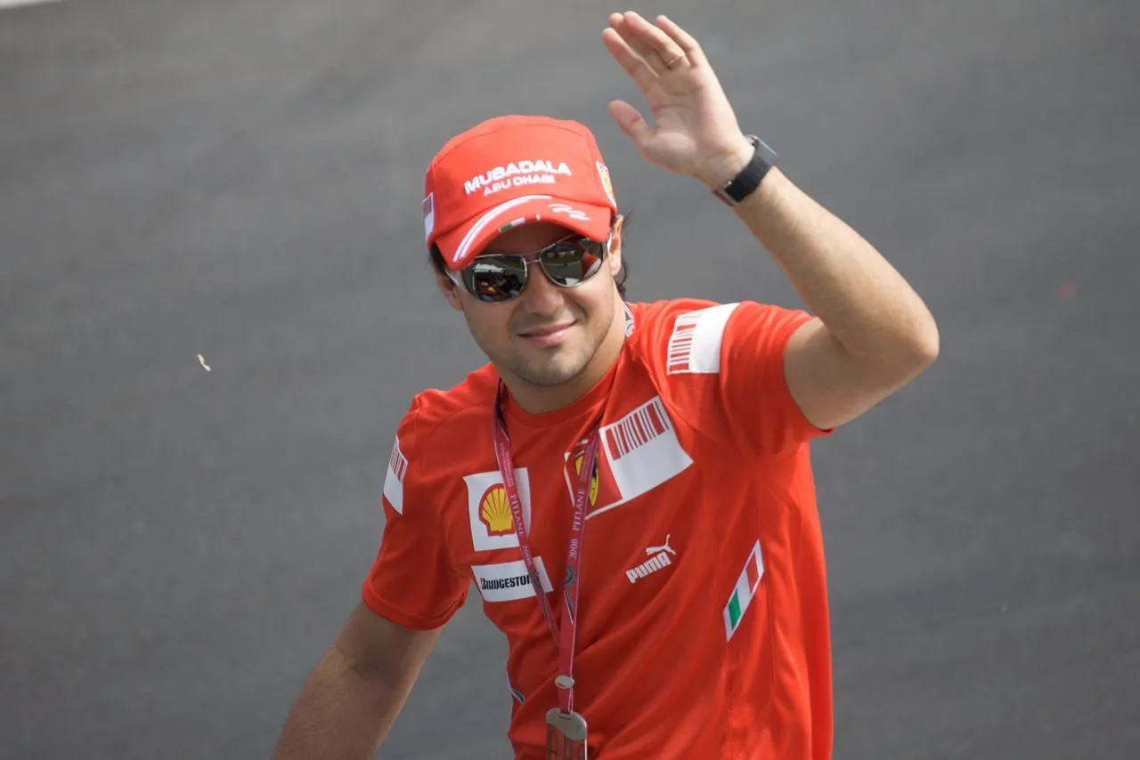 Kırmızı Ferrari takım şapkası, Shell ve Bridgestone logolu tişörtü ile boynunda 2008 Pitlane geçiş kartı bulunan Formula 1 pilotu Felipe Massa, aynalı güneş gözlükleri takılıyken gülümseyerek kameraya el sallıyor.
