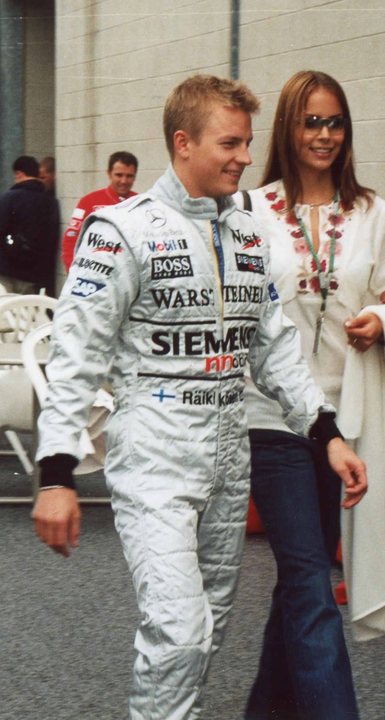 Formula 1 pilotu Kimi Räikkönen, üzerinde Mobil 1, Boss, Siemens gibi sponsor logolarının ve Finlandiya bayrağının yer aldığı gümüş renkli, kapitone yarış tulumuyla, güneş gözlüklü ve işlemeli beyaz bluz giymiş uzun saçlı bir kadının yanında neşeli bir ifadeyle yürüyor.