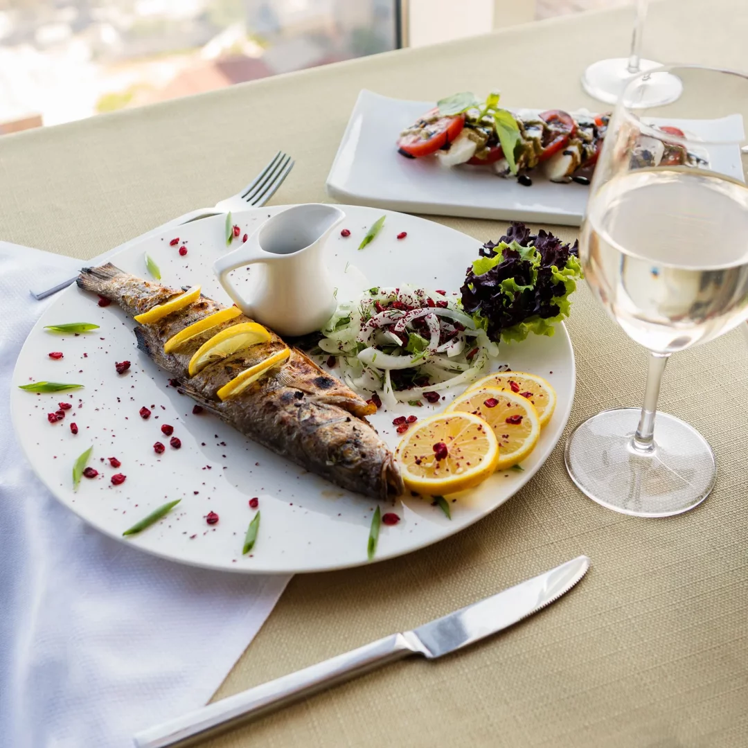 Bir restoran masasında, üzerine limon dilimleri ve sumak serpilmiş, nar ekşili soğan salatası ile birlikte sunulan bütün ızgara balık, Türk balık çeşitlerinden lezzetli bir örneği vurgularken yanında bir kadeh beyaz şarap ve arka planda başka bir salata görülmektedir.