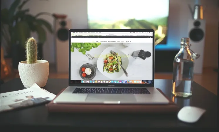 Modern bir salonda, dizüstü bilgisayar ekranında bir sağlıklı yemek tarifi web sitesi açık; yanında cam şişe ve saksı bitkisi yer alıyor.