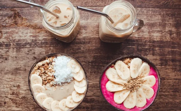 Ahşap masa üzerinde, muz dilimleriyle süslenmiş iki smoothie bowl ve cam kavanozlarda soğuk kahveler; taze ve sağlıklı bir kahvaltı.