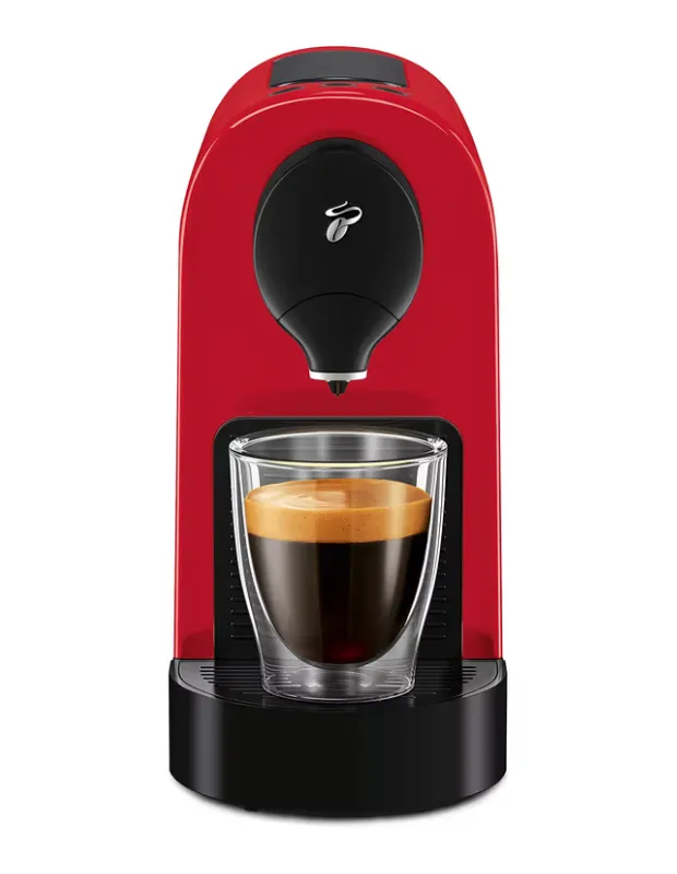 Parlak kırmızı Tchibo kapsül kahve makinesi, çift cidarlı cam bardağa taze demlenmiş, bol kremalı bir espresso dolduruyor.