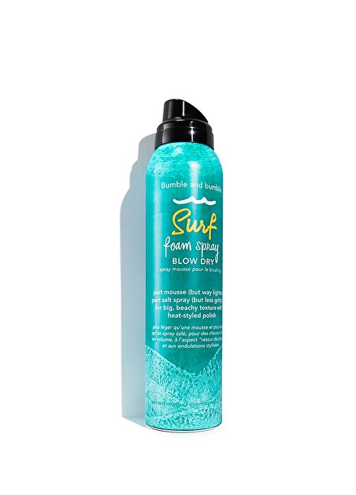 Beyaz bir zeminde dikey olarak duran, üzerinde dalga motifleri ve marka adı Bumble and bumble ile Surf foam spray BLOW DRY yazan, siyah kapaklı parlak turkuaz renkli bir şişe, fön sonrası saçlara hacim veren ve belirgin plaj dalgaları dokusu sağlayan bir saç ürününü gösteriyor.