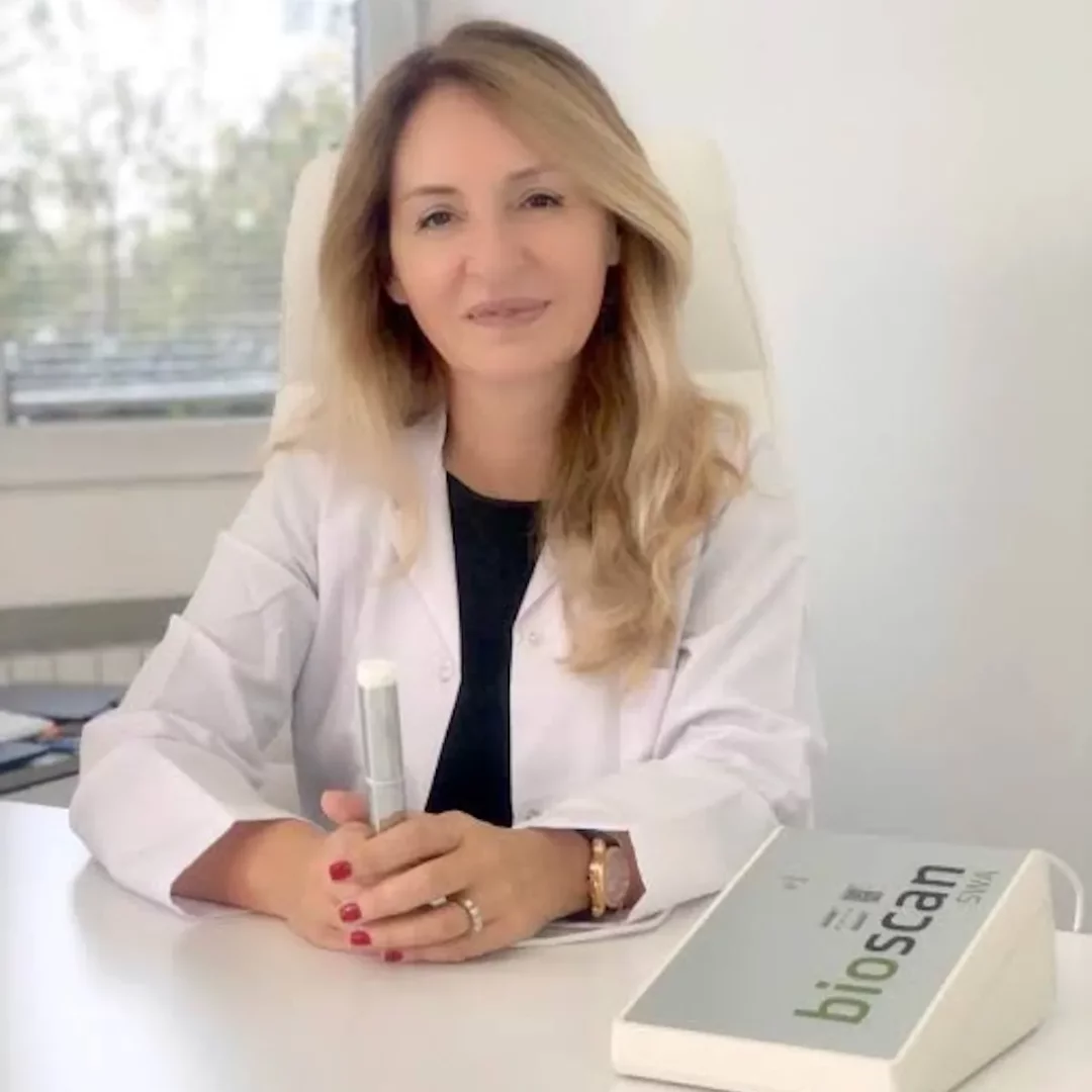 Pencere önünde, beyaz önlüğüyle gülümseyen Dr. Elçin Yıldıran Mızrahi, kliniğinde masasında oturmuş, elinde metalik bir ölçüm cihazı tutarken, yanında biyorezonans analizi için kullanılan bioscan SWA cihazı net bir şekilde görülüyor.