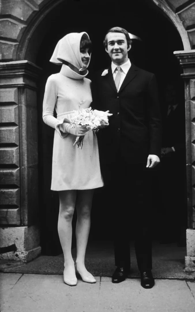 Audrey Hepburn, yüksek yakalı, uzun kollu, diz üstü minimalist Givenchy gelinliği ve uyumlu başörtüsüyle, kemerli taş bir kapının önünde, takım elbiseli eşiyle birlikte gülümsüyor, bu kare moda tarihinin en ikonik anlarından birini yansıtıyor.