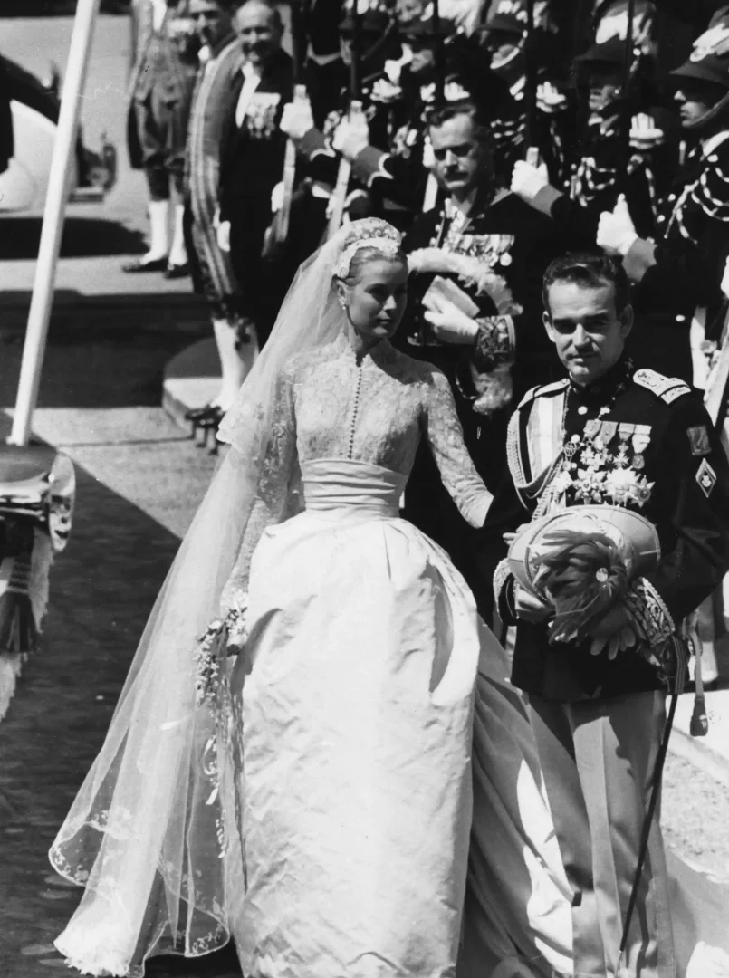 Siyah beyaz bu fotoğrafta, Helen Rose tasarımı dantel üst kısmı, bele oturan kuşağı ve kabarık ipek eteğiyle moda tarihine damgasını vuran zamansız zarafete sahip Grace Kelly gelinliği içinde Monako Prensi Rainier III ile birlikte merdivenlerden inerken görülüyorlar.