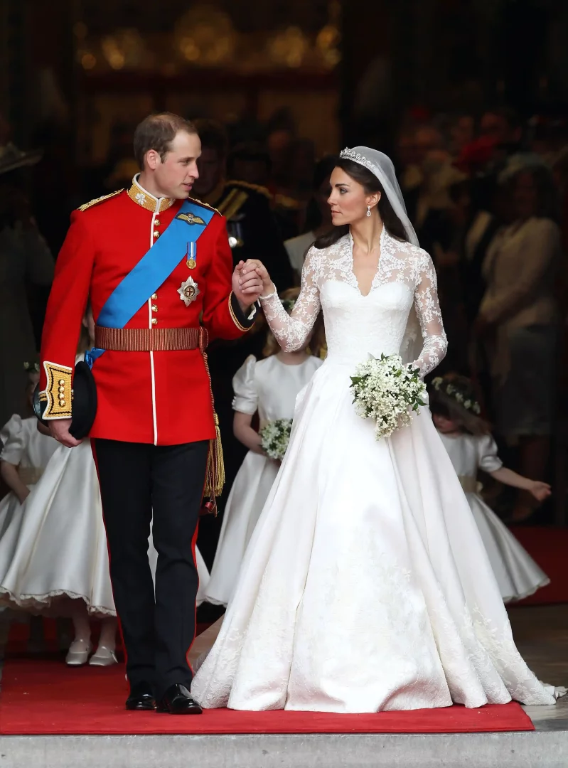 Kate Middleton ve Prens William, Kate'in Alexander McQueen tasarımı uzun kollu, dantel detaylı, derin V yakalı ikonik gelinliği ve William'ın kırmızı askeri üniforması içinde el ele tutuşarak düğünlerinden sonra kırmızı halıda ilerlerken, arkalarında beyaz elbiseli çiçek kızlar belirginleşiyor.