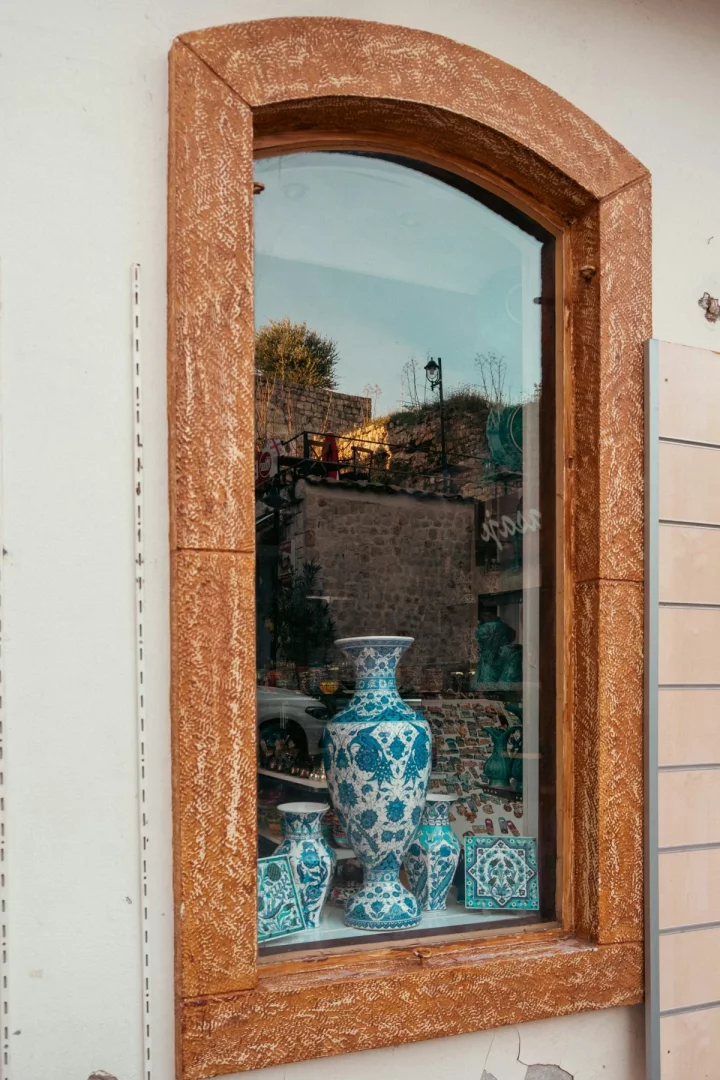 Alaçatı'daki geleneksel bir evin penceresinin ardında, karmaşık mavi-beyaz çiçek ve kuş desenleriyle bezeli gösterişli bir seramik vazo, çevresindeki küçük vazolar ve el yapımı karolarla birlikte şık bir dekorasyon detayı sunuyor, camda dış mekanın yansıması beliriyor.