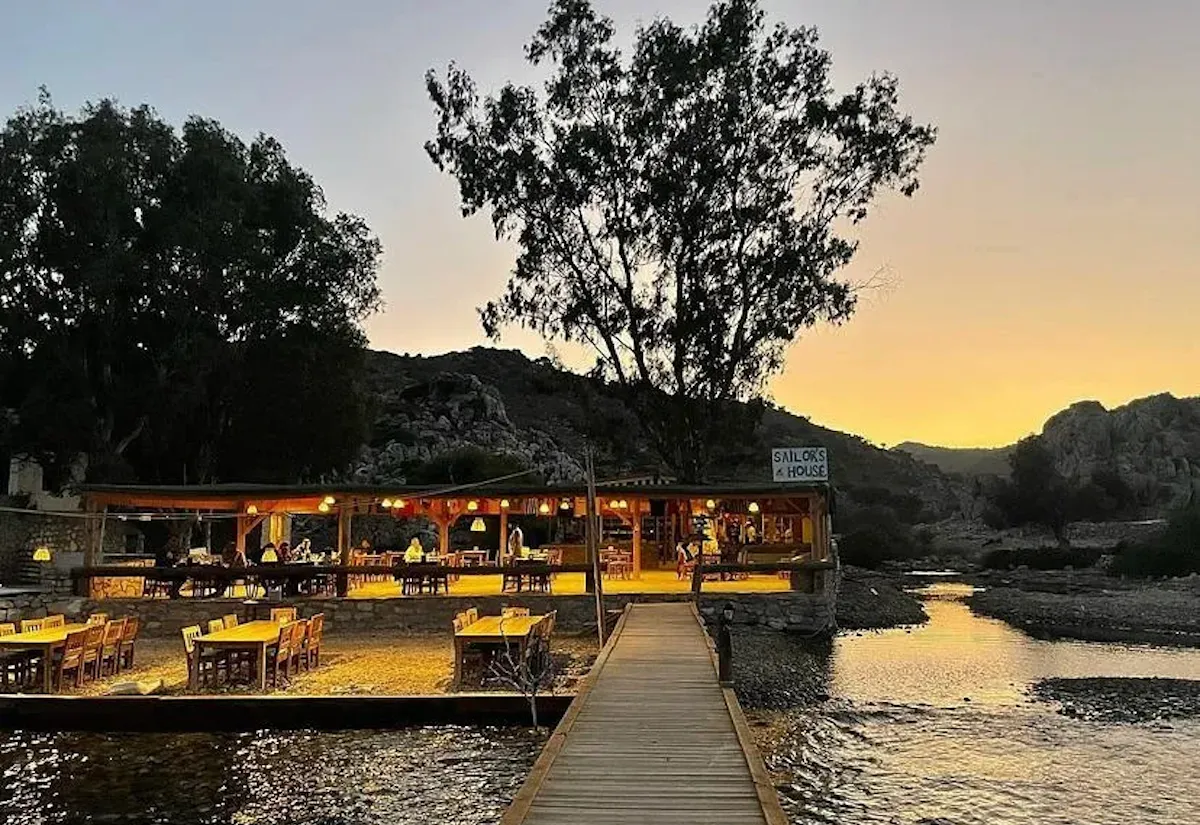 Alacakaranlıkta, Marmaris'in Bozukkale bölgesinde, sakin su üzerinde uzanan ahşap iskele ile sıcak ışıklarla aydınlatılmış açık ve kapalı alanlarıyla misafirlerini ağırlayan Sailor's House restoranının keyifli bir akşam manzarası.