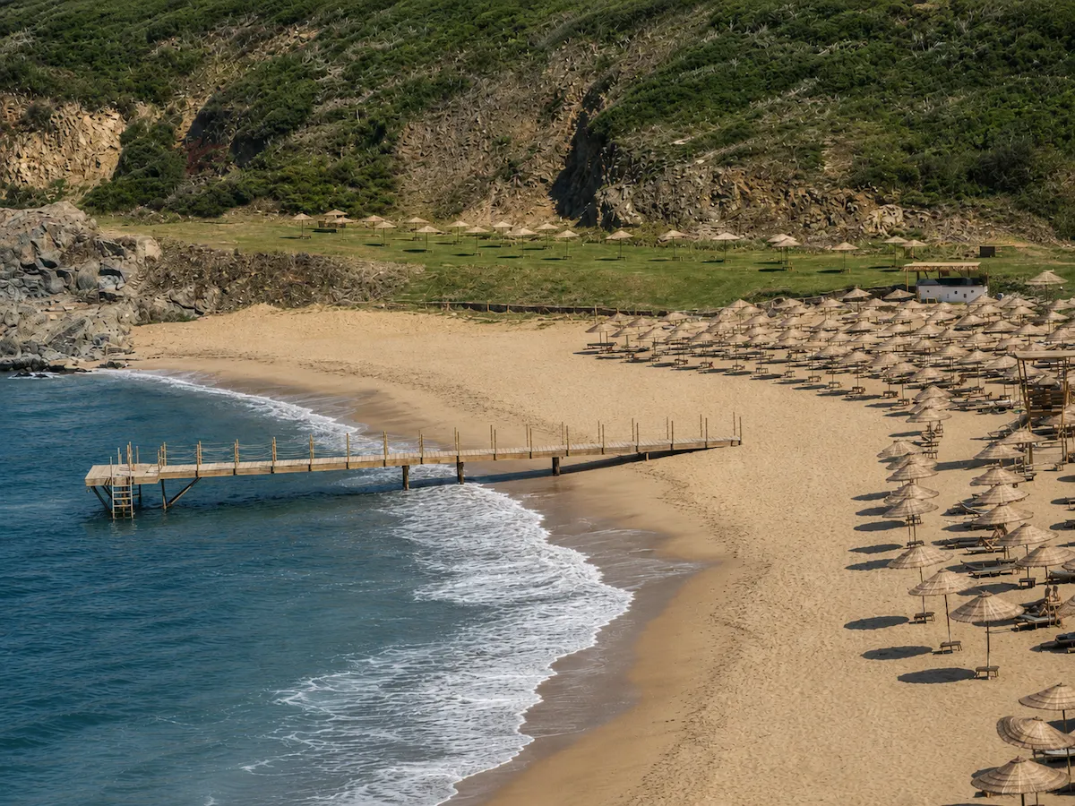 Kilyos Milyon Beach'te, berrak mavi denize doğru uzanan ahşap bir iskele, dalgaların vurduğu altın rengi kumlu sahil boyunca düzenli sıralar halinde dizilmiş yüzlerce hasır şemsiye ve şezlong ile yemyeşil kayalık tepelerin kucakladığı sakin bir koy manzarası.