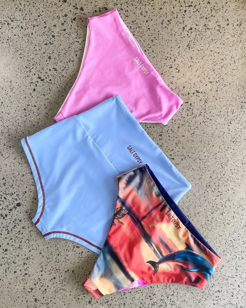 Beton zemin üzerinde yan yana duran, Salt Gypsy markasına ait pembe, açık mavi ve yunus ile kaplumbağa desenli, parlak renkli üç farklı bikini altı, sürdürülebilir mayo ve bikini seçeneklerini öne çıkarıyor.