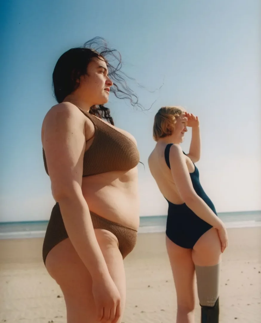 Sahilde, esen rüzgarda saçları dalgalanan, kahverengi ribanalı bikini giymiş kıvrımlı bir kadın ve arkasında lacivert tek parça mayosuyla güneşe bakarken protez bacağı görünen sarı saçlı bir kadın, sürdürülebilir mayo ve bikini markalarının çeşitliliğini yansıtıyor.