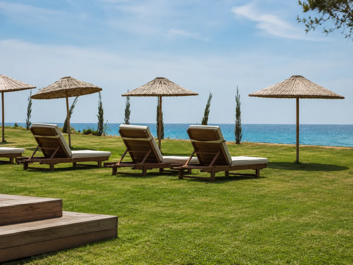 Çeşme Plajı'nda, yemyeşil çimenler üzerinde sıralanmış hasır şemsiyelerin gölgesindeki açık renk minderli ahşap şezlonglar, Ege Denizi'nin masmavi sularına bakan huzurlu bir dinlenme alanı sunuyor.