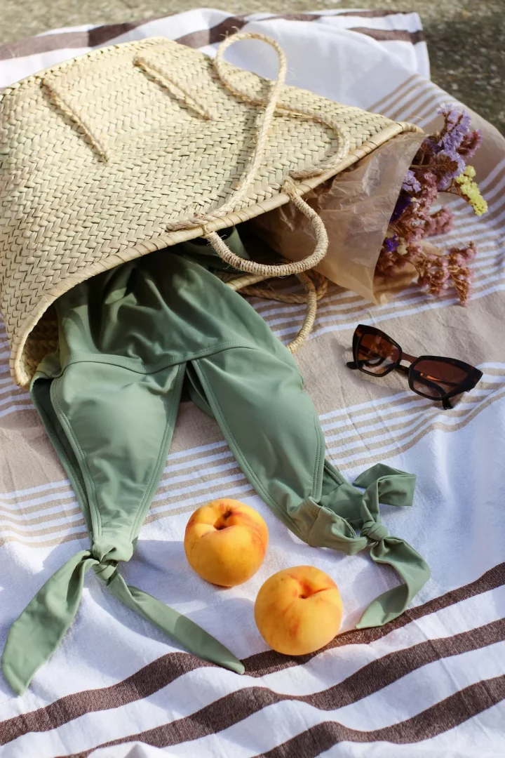 Beyaz ve kahverengi çizgili bir havlu üzerinde, içinden yeşil bir bikini üstü ve kahverengi kağıda sarılı renkli kuru çiçeklerin sarktığı hasır bir plaj çantası, yanında duran şık güneş gözlüğü ve iki olgun şeftaliyle yazın tadını çıkarılacak bir anı yansıtıyor.