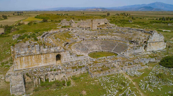 Milet Antik Kenti'nin geniş bir alana yayılan, basamaklı oturma sıralarına ve merkezi arenaya sahip, Helenistik ve Roma dönemlerinden kalma iyi korunmuş taş tiyatrosu, yemyeşil otlar ve uzaktaki tarım arazileriyle çevrili bir manzara içinde görülüyor.