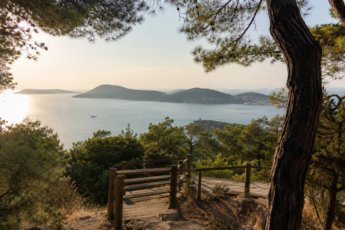 İstanbul Büyükada ekoturizm alanından çekilen bu görselde, çam ağaçlarının çevrelediği toprak bir patikadan ahşap korkuluklu bir seyir terasının ötesindeki Marmara Denizi'nin ve güneşin yansıdığı ufukta uzanan adaların büyüleyici manzarası yansıyor.