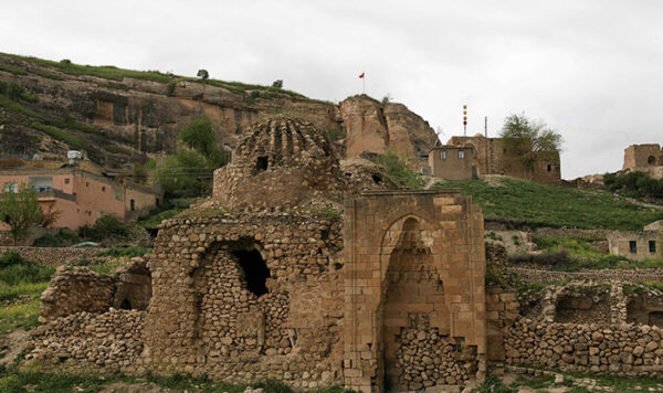 Gri, bulutlu bir gökyüzünün altında, Hasankeyf Antik Kenti'nin engebeli yamaçlarında yer alan, kısmen harabe haldeki taş yapılar, geleneksel evler ve zirvedeki kale kalıntıları yemyeşil bitki örtüsüyle bezenmiş bir tarih manzarasını gözler önüne seriyor.