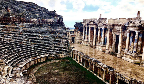 Islak zemini ve kısmen bulutlu gökyüzü altında, solda yükselen basamaklı oturma düzeni ve sağda detaylı oyulmuş sütunlu sahne binasıyla Hierapolis Antik Kenti'nin görkemli tiyatrosu, Türkiye'nin antik kentleri rehberinde öne çıkan büyüleyici tarihi yapılarından biridir.
