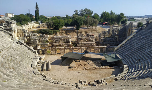 Myra Antik Kenti'nin iyi korunmuş taş basamaklı tiyatrosu, sahne alanındaki devam eden kazı çalışmaları ve kalıntılarıyla, arka planda ağaçlar ve uzaklardaki modern yapılarla çevrili bir manzarayı gözler önüne sererek Türkiye'nin zengin tarihi antik kentler mirasını yansıtıyor.