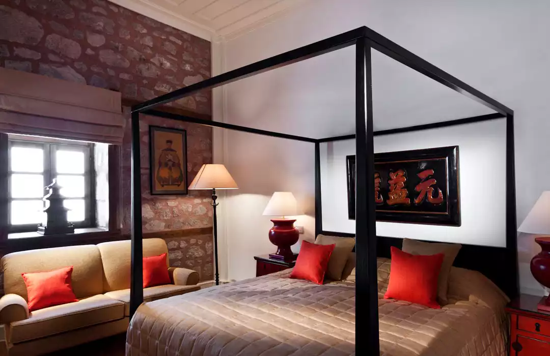 Divan Hotels'ın otantik mimarisine sahip lüks bir odasında, tavandan yere uzanan siyah direkli geniş yatak, kırmızı yastıklı bej bir divan ve taş duvarlarla çevrili şık atmosferde, Uzak Doğu motifli tablolar ve yumuşak ışıklarla özel bir konaklama deneyimi sunulmaktadır.