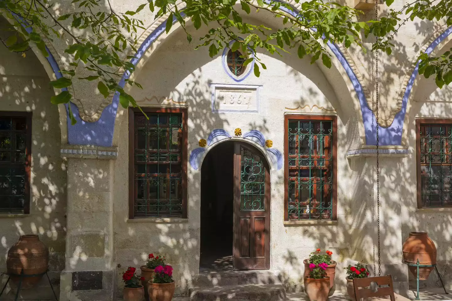 Tarihi Gül Konakları'ndan birinin 1864 tarihli, kemerli ahşap kapısı ve demir parmaklıklı pencereleriyle süslü bej renkli girişi, asma yapraklarının gölgelerinin düştüğü duvarları ve canlı kırmızı-pembe çiçeklerle dolu saksılarıyla sıcak bir atmosfer sunuyor.