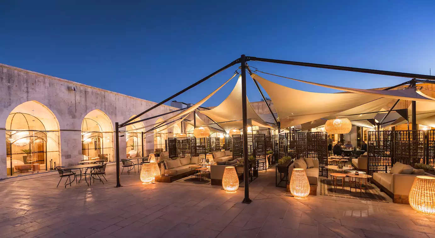 Akşam alacakaranlığında, geniş açık hava mekanında yer alan şık bir restoran veya lounge alanı, beyazımsı kumaş tentelerin altında, kemerli taş duvarların önünde, hasır detaylı aydınlatmalarla sıcak bir ambiyans yaratan konforlu oturma grupları ve masalarıyla misafirlerini bekliyor.