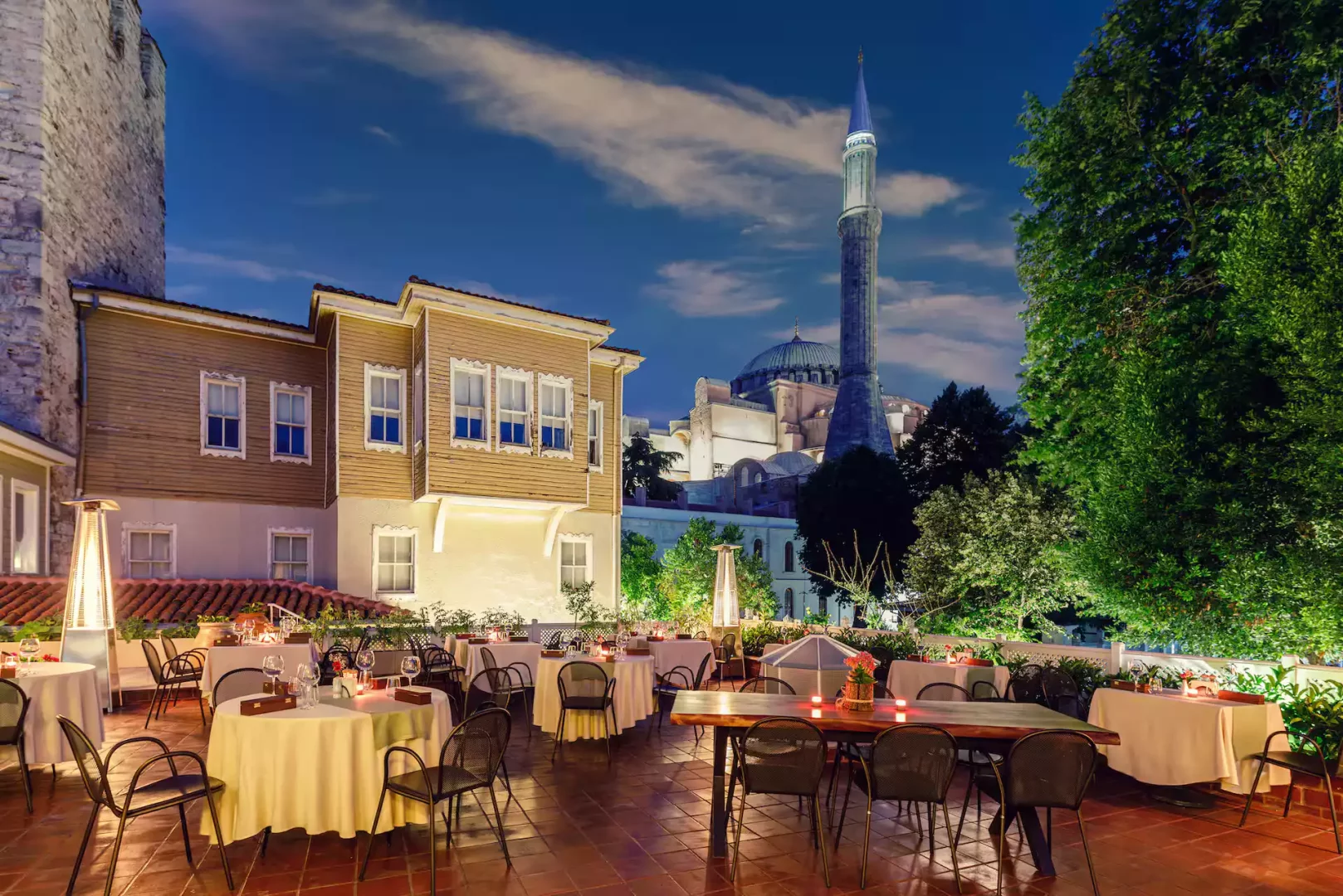 Akşam karanlığında, geleneksel ahşap bir evin önündeki şık açık hava teras restoranında, masa düzenlemeleri ve ısıtıcı lambalarla donatılmış zeminde, arka planda Ayasofya'nın kubbesi ve zarif minaresi göz alıcı bir şekilde aydınlatılmış.