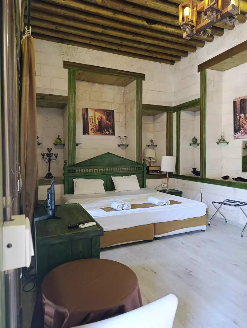 Bir butik otel odası, taş duvarları, ahşap tavan kirişleri ve yeşil detaylı ahşap mobilyalarıyla otantik bir atmosfer sunarken, geniş yatakta katlanmış havlular ve modern bir televizyon dikkat çekiyor.