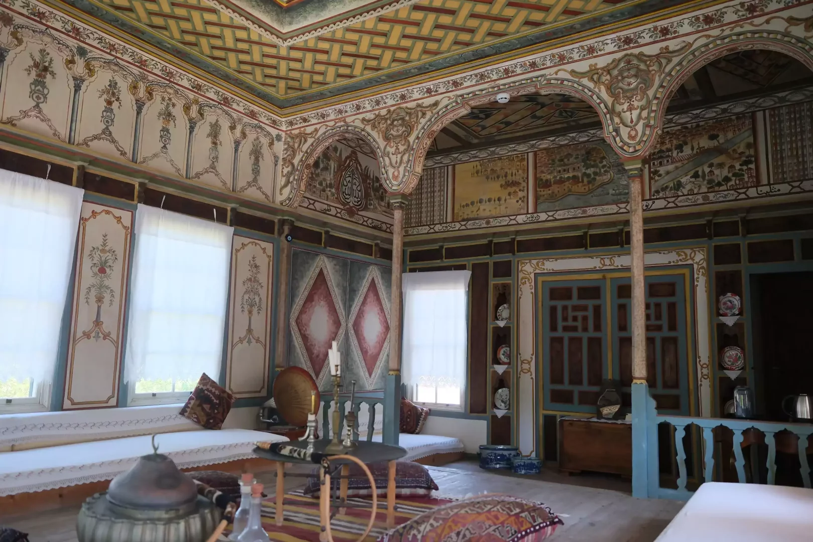 Geleneksel Türk mimarisinin zenginliğini sergileyen bu görkemli odada, geometrik desenlerle süslü ahşap tavan, çiçek ve manzara motifli duvar freskleri, zarif kemerler, pencereleri örten beyaz tüller ve etrafı minderlerle donatılmış sedirler dikkat çekerek kültürel mirasın derinliğini vurguluyor.