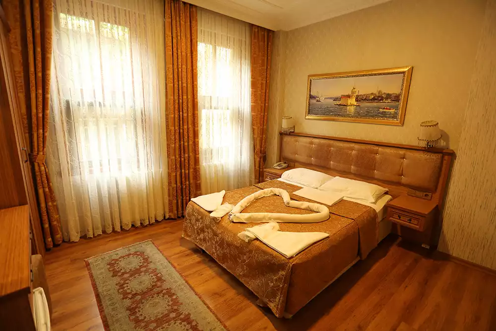 Hotel Tashkonak'ın misafirperverliğini yansıtan, altın rengi desenli çift kişilik yatakta beyaz havlulardan oluşturulmuş kalp şekli ve pencereden süzülen ışıkla aydınlanan, ahşap zeminli, İstanbul manzaralı tabloyla dekore edilmiş konforlu bir otel odası.
