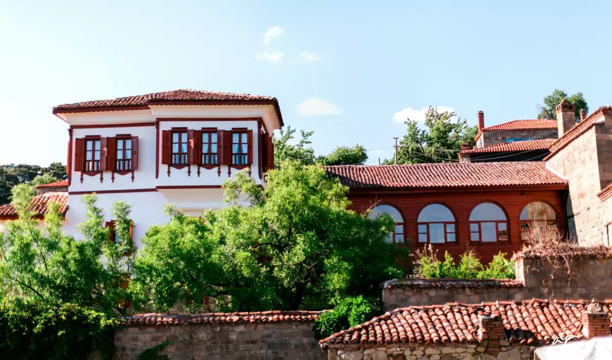 Hunnap Han'ın beyaz sıvalı dış cephesi, kahverengi ahşap pencereleri ve kırmızı kiremit çatısıyla öne çıktığı, etrafını saran yemyeşil ağaçlar ve diğer geleneksel Türk mimarisi örnekleriyle çevrili tarihi bir mahalle manzarası.