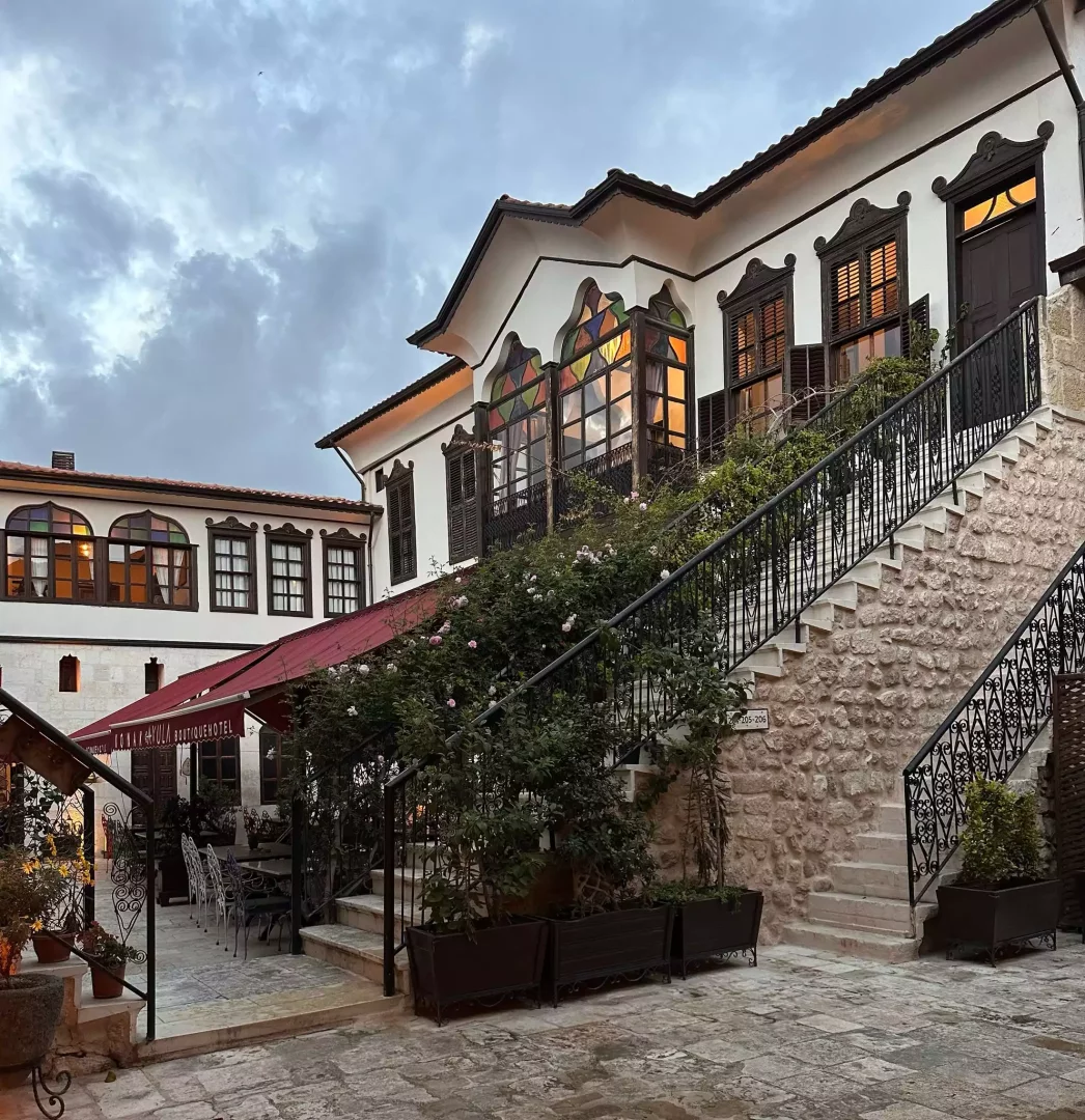 Beyaz sıvalı duvarları, ahşap kepenkleri ve renkli vitray pencereleriyle geleneksel bir mimariye sahip Konak Avula Butik Otel'in taş avlusunda, etrafı güllerle süslü taş merdivenler ve kırmızı tenteli dış mekan oturma alanı akşamüstü ışığında huzurlu bir görünüm sunuyor.