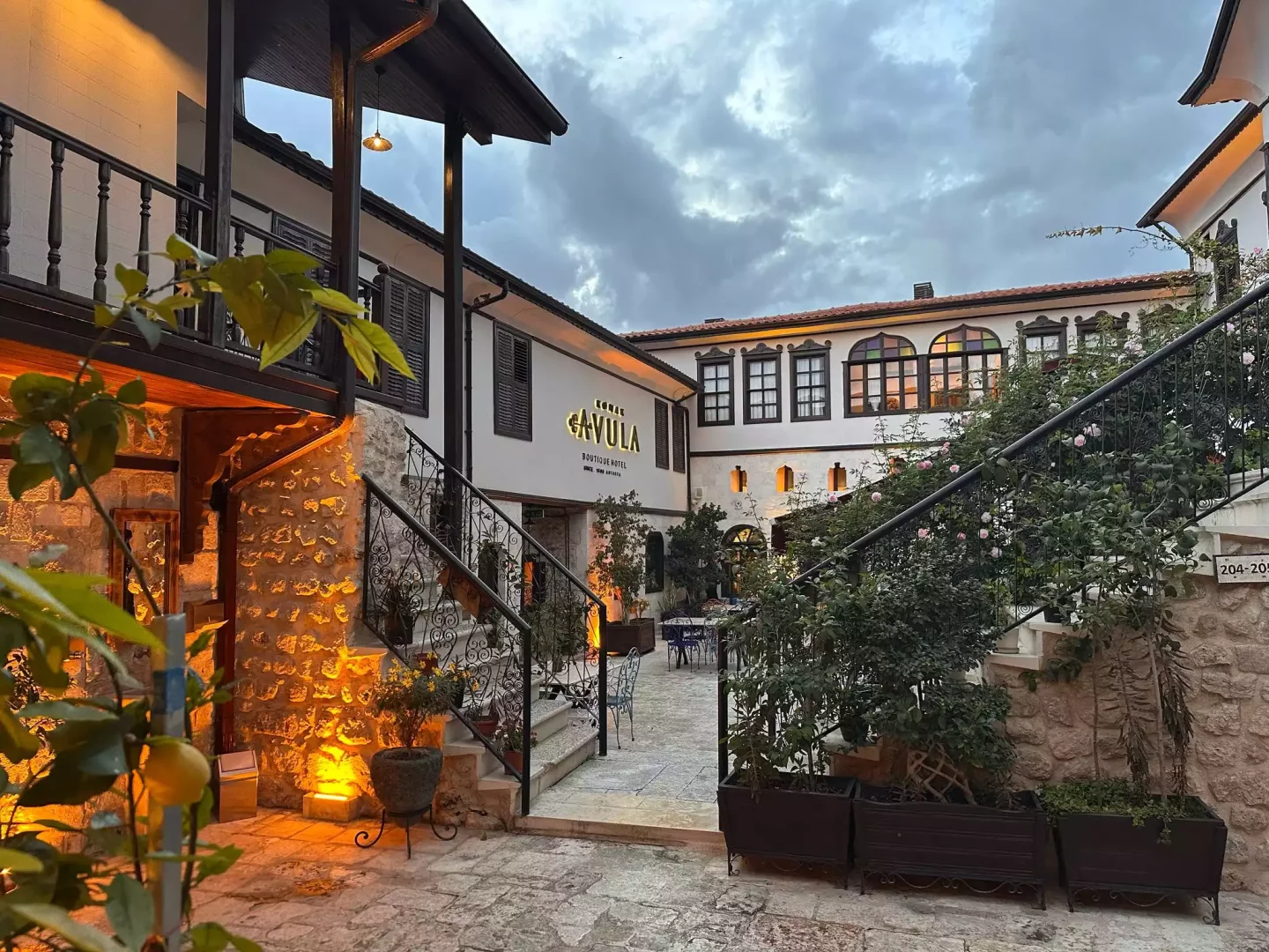 Konak Avula Boutique Hotel'in taş döşeli avlusunda, geleneksel beyaz binaların ahşap detayları ve taş duvarlar sıcak ışıklarla aydınlanırken, demir korkuluklu merdivenleri saran güllü bitkiler ve ön plandaki limon ağacı, bulutlu akşamüstünde samimi bir atmosfer sunuyor.