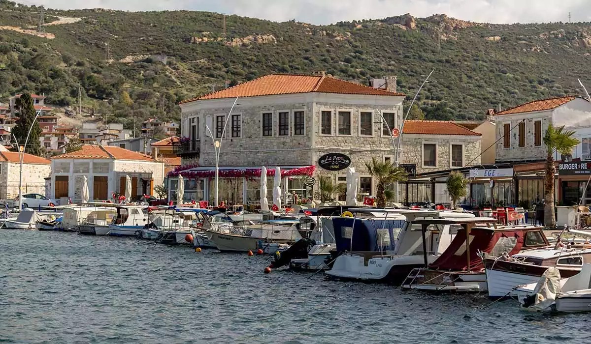 Mavi denizde sıralanmış teknelerin önünde, geleneksel taş mimarili La Petra Hotel ve çevresindeki restoranların kırmızı kiremitli çatılarıyla öne çıktığı, yeşil tepelerle çevrili şirin bir Ege kıyı kasabasının hareketli limanı.