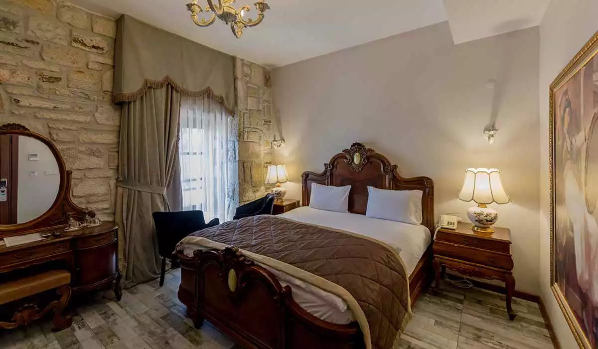 Oymalı koyu ahşap karyola ve komodinlerin eşlik ettiği şık bir otel odasında, sol duvarda doğal taş dokusu dikkat çekerken, pencereden süzülen ışık bej tonlarındaki kalın perdelerle yumuşatılıyor; odanın ortasında kahverengi kapitone yorgan örtülü, beyaz yastıklı büyük bir yatak yer alıyor.