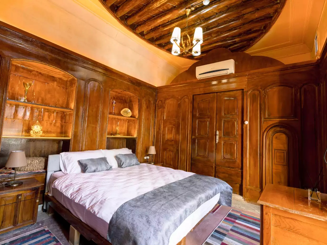 Geleneksel ahşap panellerle kaplı duvarlara ve özel tasarım ahşap tavan işlemeciliğine sahip, beyaz çarşaflı ve gri yastıklı konforlu bir çift kişilik yatağın bulunduğu, lüks dekorasyonlu ve zarif avizeli ferah bir yatak odası.