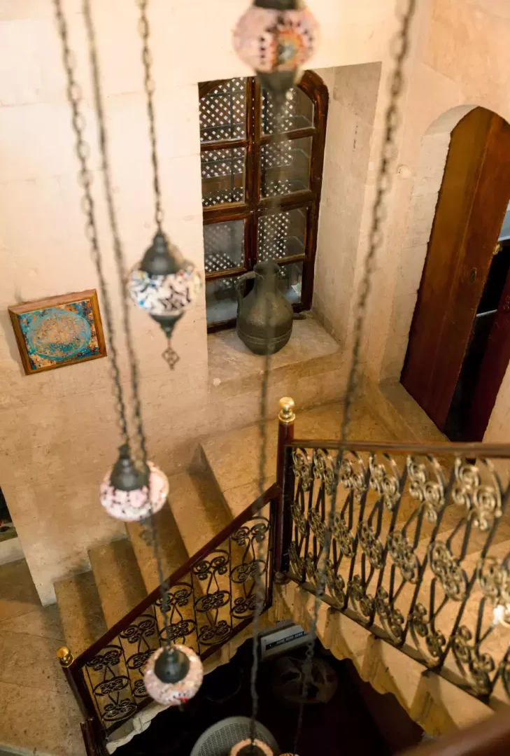 Yukarıdan aşağıya doğru çekilmiş bu fotoğrafta, ahşap çerçeveli kafesli bir pencere ve pencere önünde duran eski bir sürahi ile aydınlık taş merdivenler, oymalı ferforje bir korkuluk ve tavandan zincirlerle sarkan renkli mozaik lambalar görünen geleneksel bir iç mekan.