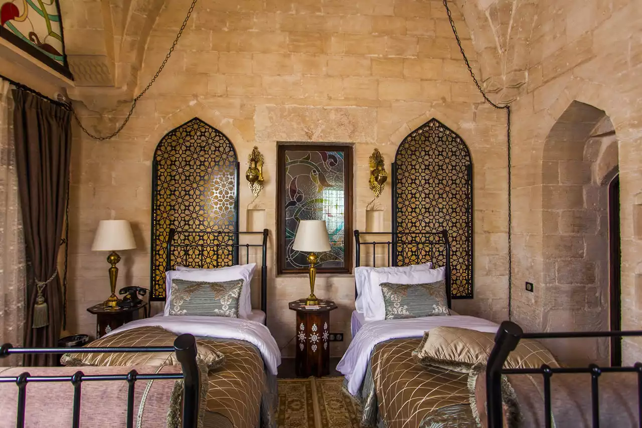 Mardin'in taş mimarisine uygun tasarlanmış otantik bir Mardius otel odasında, vitraylı pencerenin iki yanında geometrik desenli kemerli panjurların önünde yer alan iki tek kişilik yatak, pirinç tabanlı zarif lambalar ve işlemeli komodinlerle tarihi ve konforlu bir atmosfer sunuyor.
