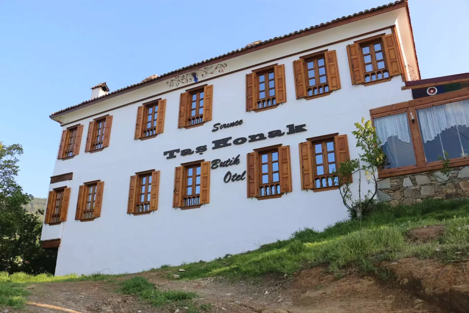 Beyaz cephesi, kahverengi ahşap pencereleri ve açık panjurları olan, üzerinde Şirince Taş Konak Butik Otel yazan, yokuş yukarı uzanan toprak ve otlu bir yoldan çekilmiş üç katlı geleneksel konak, masmavi bir gökyüzünün altında duruyor.